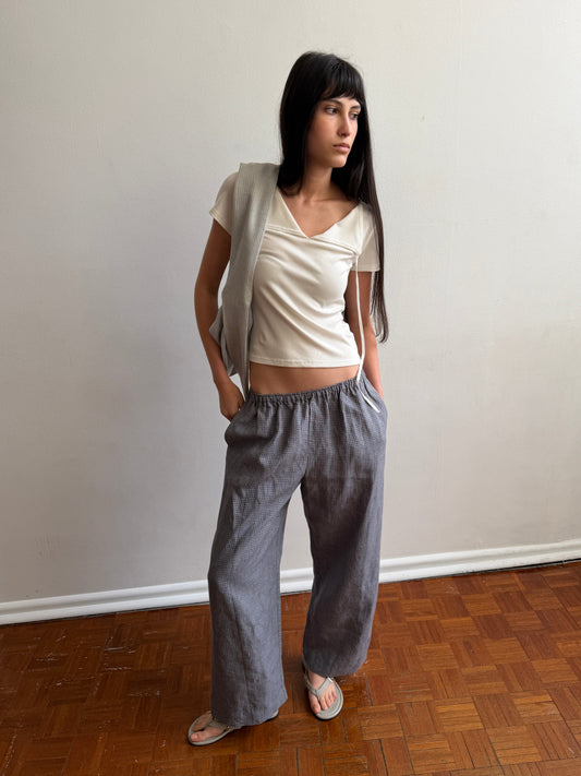A woman with dark hair and a fringe stands against a pale wall wearing the Tie Back Tee - White by Deiji Studios, a fitted cream tee with a sweetheart neckline, with grey check trousers and a cream cardigan over one shoulder.
