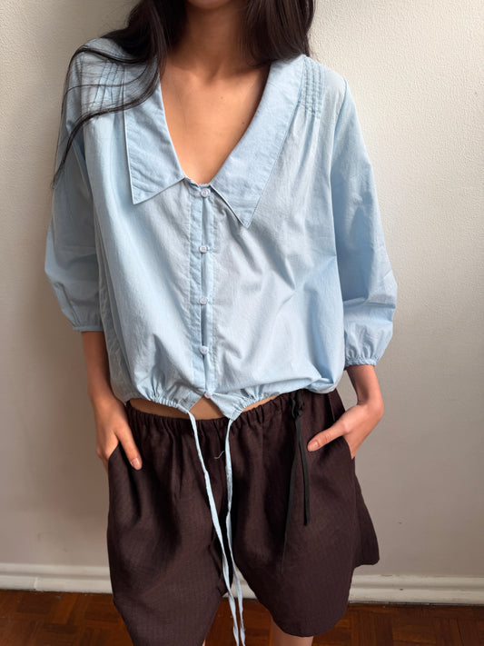 A cropped view of the Shoulder Tucks Top - Baby Blue by Deiji Studios, a baby blue blouse with a large pointed collar, buttoned V-placket and drawcord hem, worn with brown shorts against a pale wall.