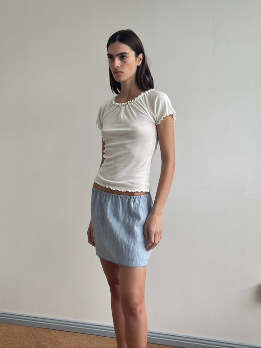 A person with straight dark hair stands indoors against a plain light wall, wearing a fitted white short-sleeve top and the Babylock Skirt - Blue Stripe. They have a neutral expression and their hands are at their sides.