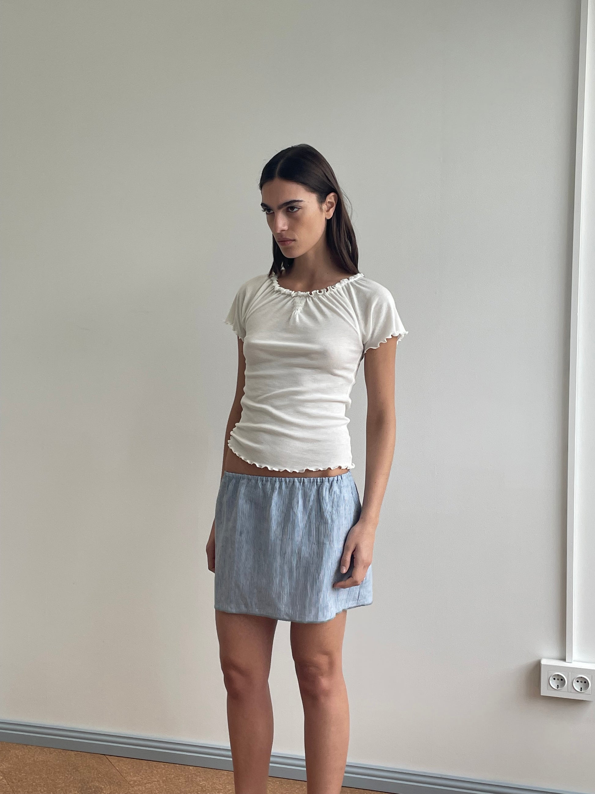 A woman with straight dark hair stands indoors against a light wall, wearing a fitted Flounce Tee - Cream by Deiji Studios and a light blue skirt. She looks slightly to the side with a neutral expression.