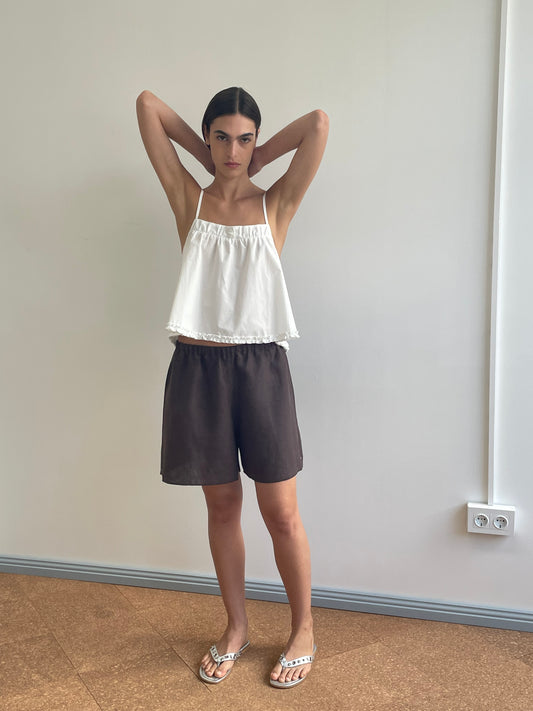 A woman stands indoors against a plain wall, wearing the Yoke Top - White by Deiji Studios, loose brown shorts, and sandals. With her arms raised and hands behind her head, she looks directly at the camera.