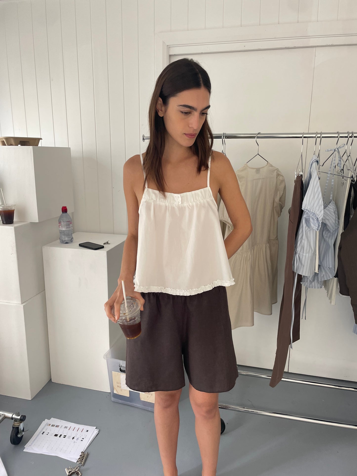 A woman in a Deiji Studios Yoke Top - White and dark shorts holds an iced coffee, standing in a minimalist room with clothes on a rack and papers scattered on the floor.
