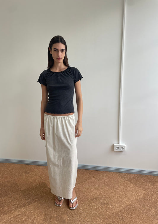 A woman stands indoors against a plain wall, wearing the Flounce Tee - Grey, a long white skirt by Deiji Studios, and sandals. She has straight dark hair and a neutral expression.