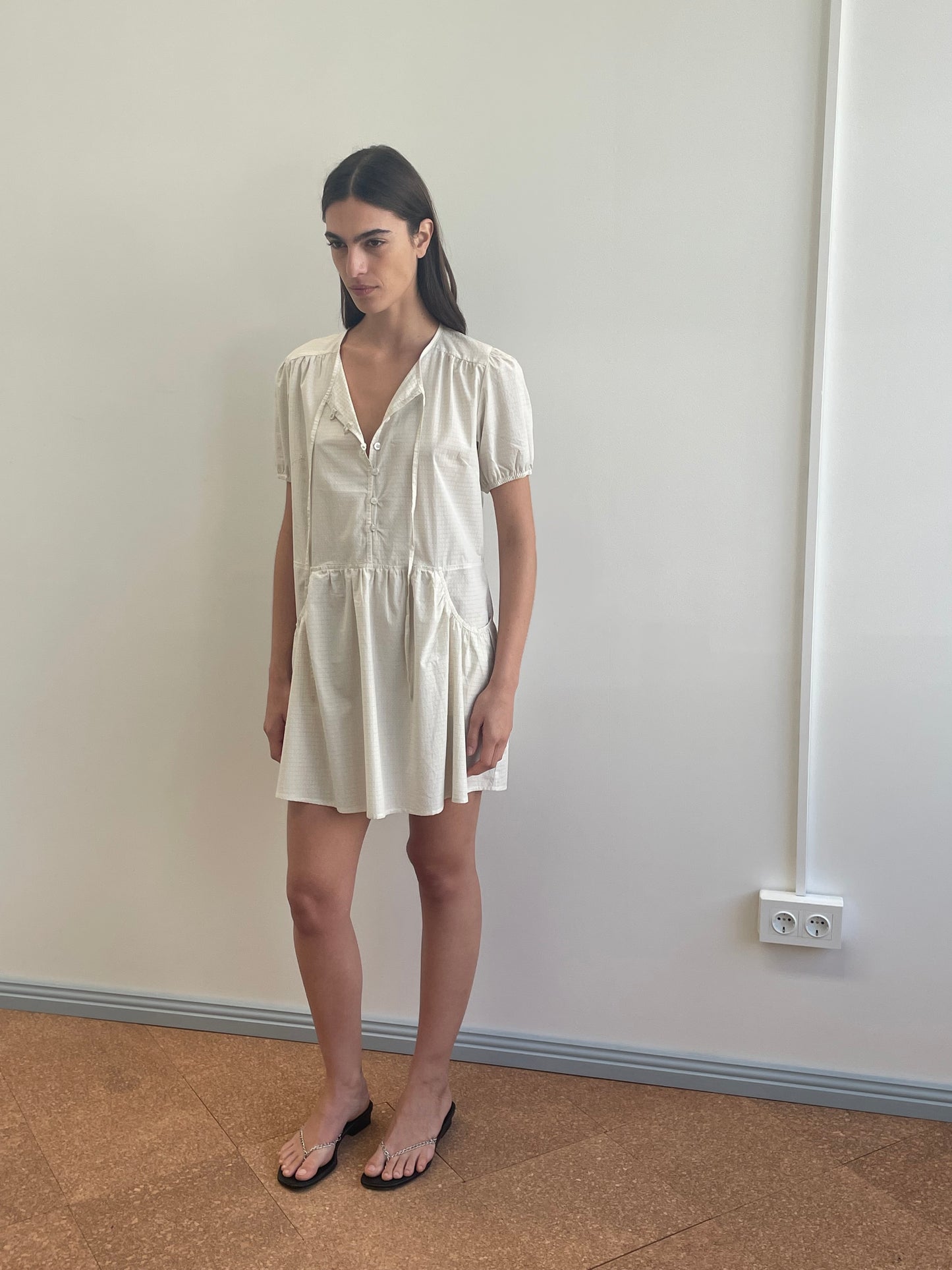 A woman with long dark hair wears the Deiji Studios Pocket Mini Dress - Bone Check and black sandals, standing indoors against a plain white wall with a power outlet near the floor.