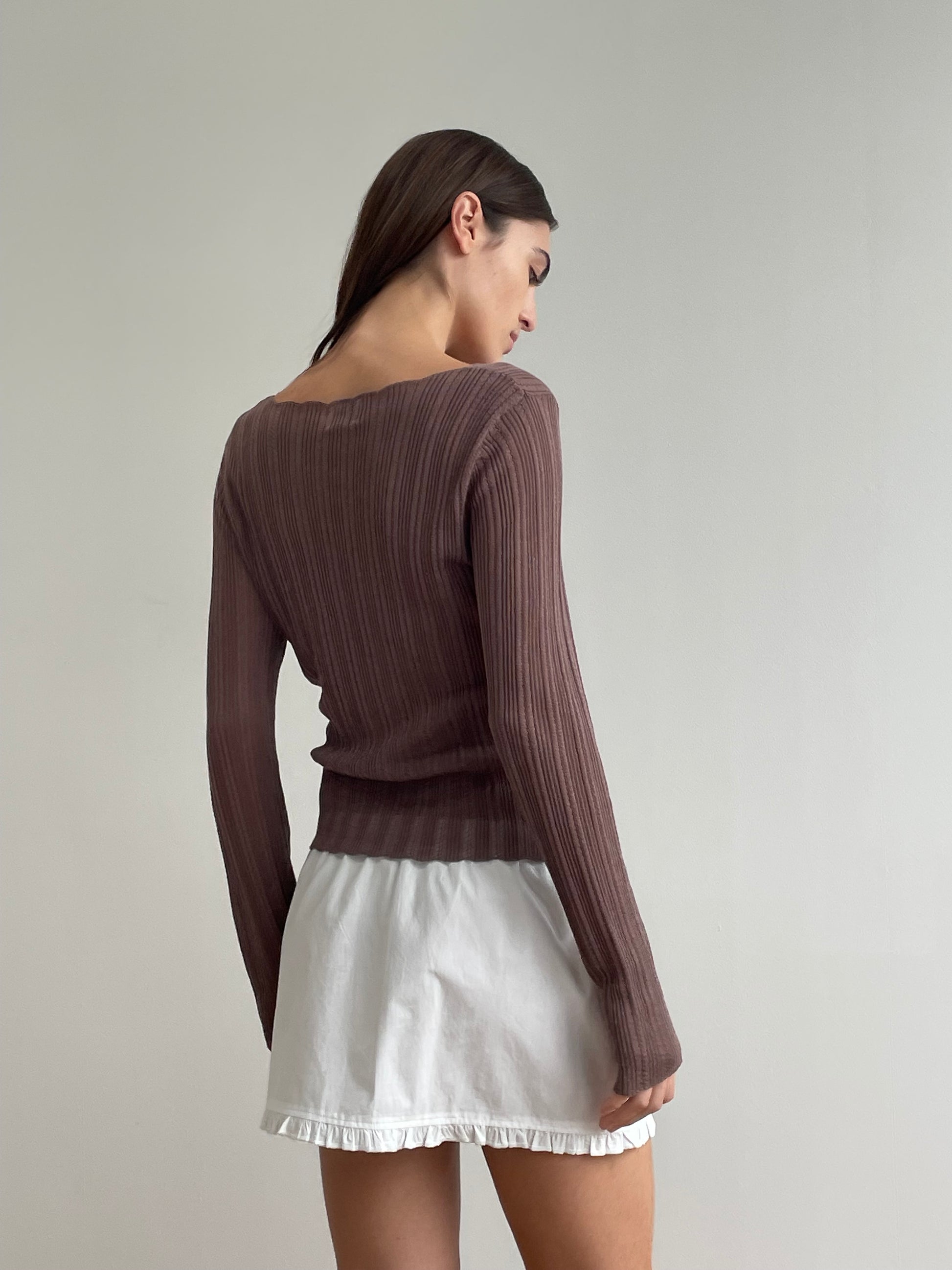 A woman with straight brown hair, facing away from the camera against a plain light wall, wears a long-sleeved ribbed mauve top and the Flounce Mini - White, an organic cotton mini skirt featuring a ruffled hem.