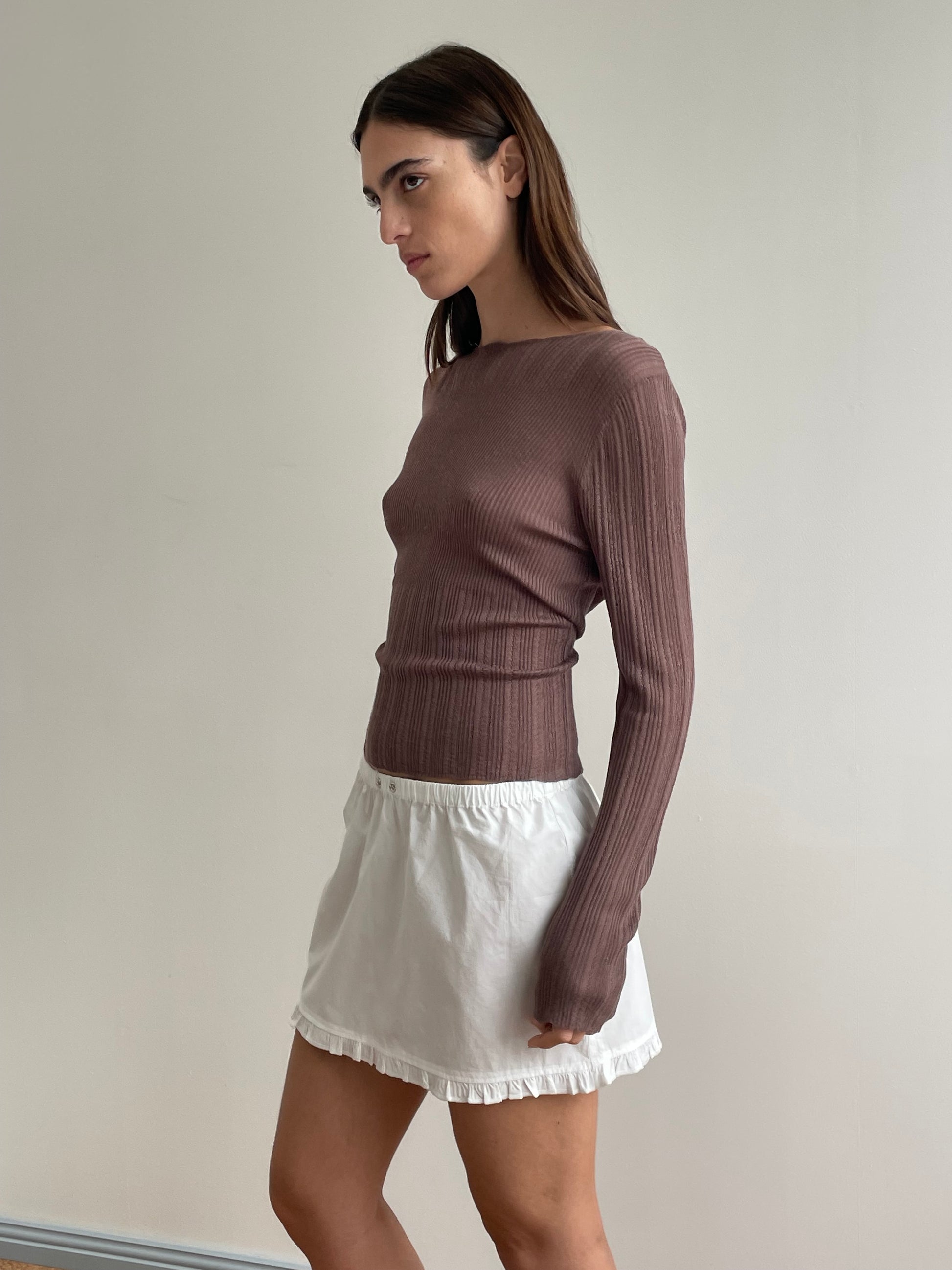 A woman with long brown hair wears a ribbed, long-sleeve mauve top and the Flounce Mini - White by Deiji Studios, featuring a ruffled hem, as she stands against a plain light-colored wall.