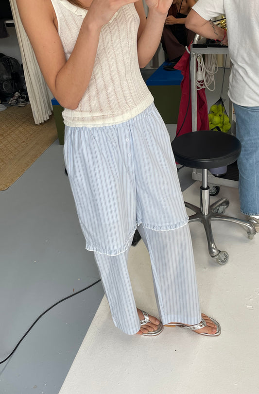 A person wears the Flounce Short - Mineral Stripe sleeveless knitted top with loose striped pants and sandals, standing indoors on a light floor. Only their upper body and legs are visible, with people and office items in the background.