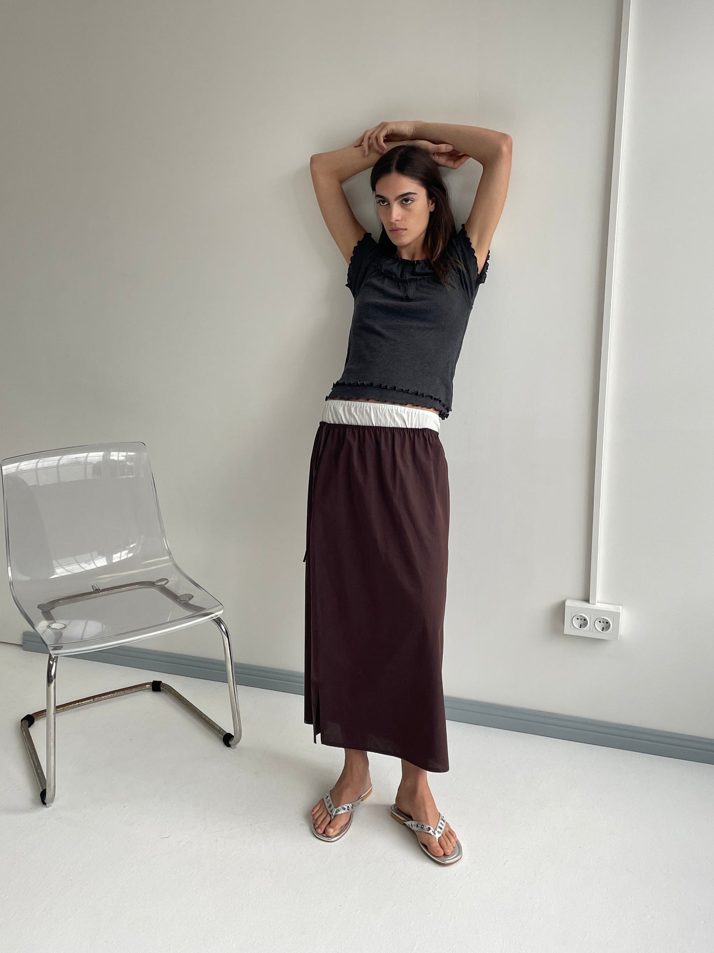 A woman stands indoors by a clear chair, wearing a black t-shirt and the Deiji Studios Mid Day Skirt - Umber with a white waistband. She raises her arms behind her head, looking at the camera in a pale room with a power outlet.