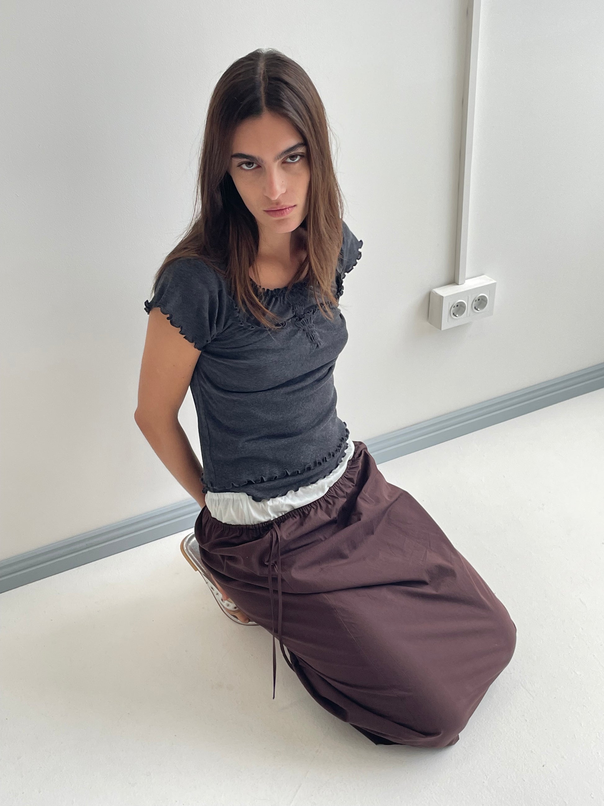 A woman with long brown hair kneels on a white floor, hands behind her back, wearing a dark t-shirt and the Mid Day Skirt - Umber. She gazes seriously at the camera; a wall with electrical outlets is behind her.
