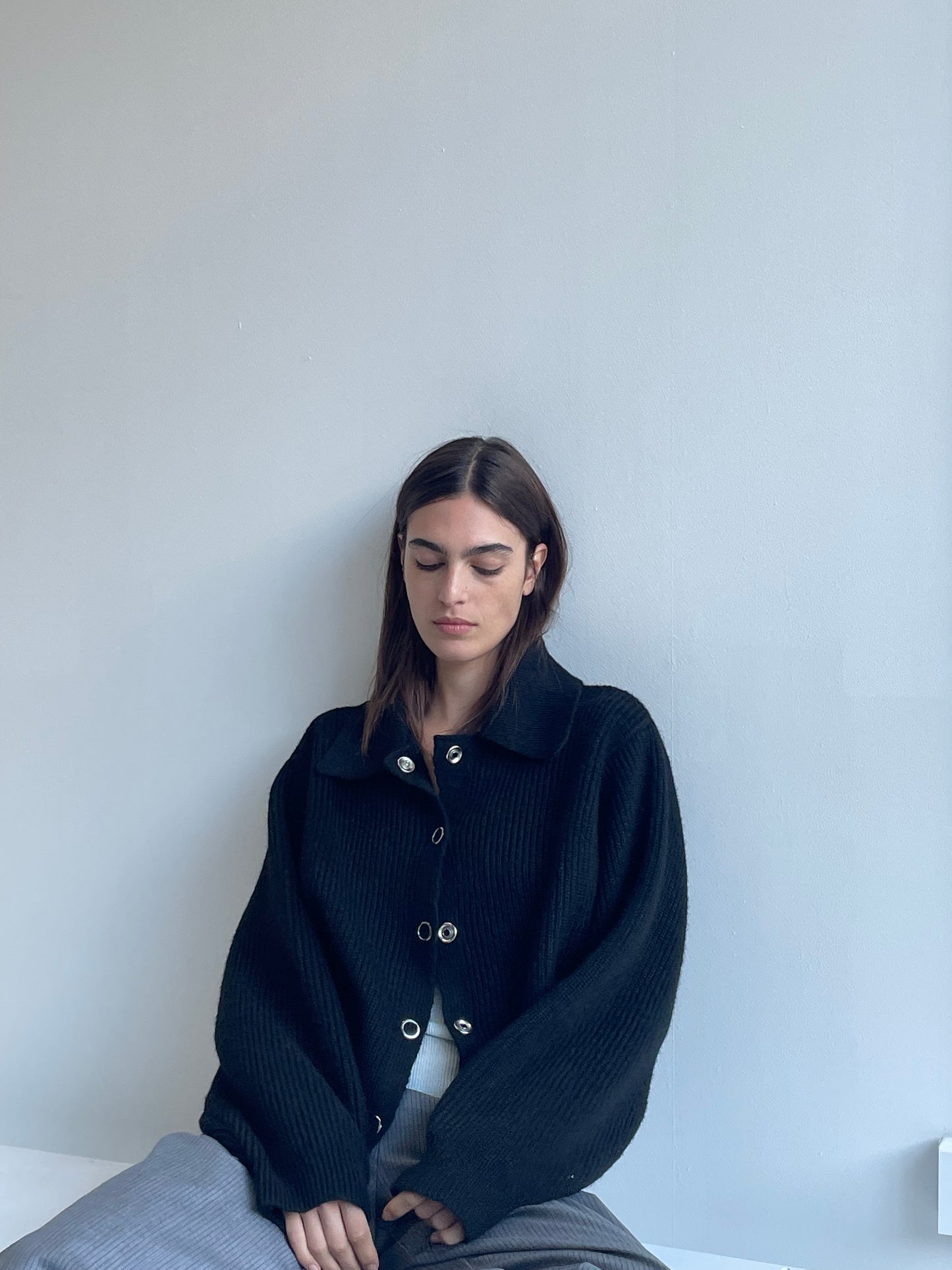 A woman with long brown hair sits against a light gray wall, wearing the Stud Cardi Knit - Black in an oversized fit and gray pants. She looks down, her expression calm and serene.