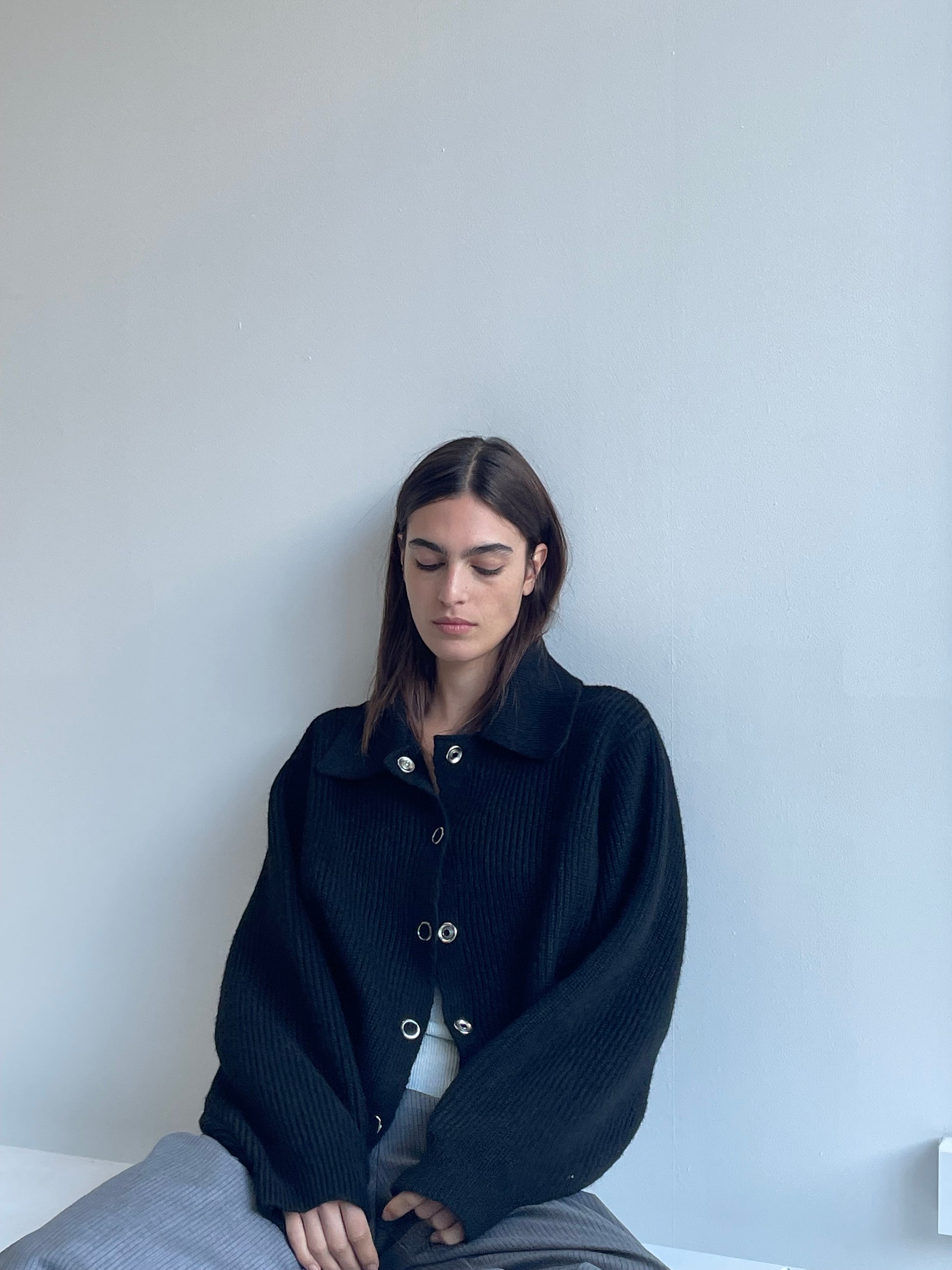 A woman with long brown hair sits against a light gray wall, wearing the Stud Cardi Knit - Black in an oversized fit and gray pants. She looks down, her expression calm and serene.