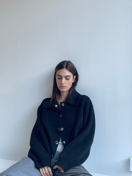 A woman with long brown hair sits against a light gray wall, wearing the Stud Cardi Knit - Black in an oversized fit and gray pants. She looks down, her expression calm and serene.