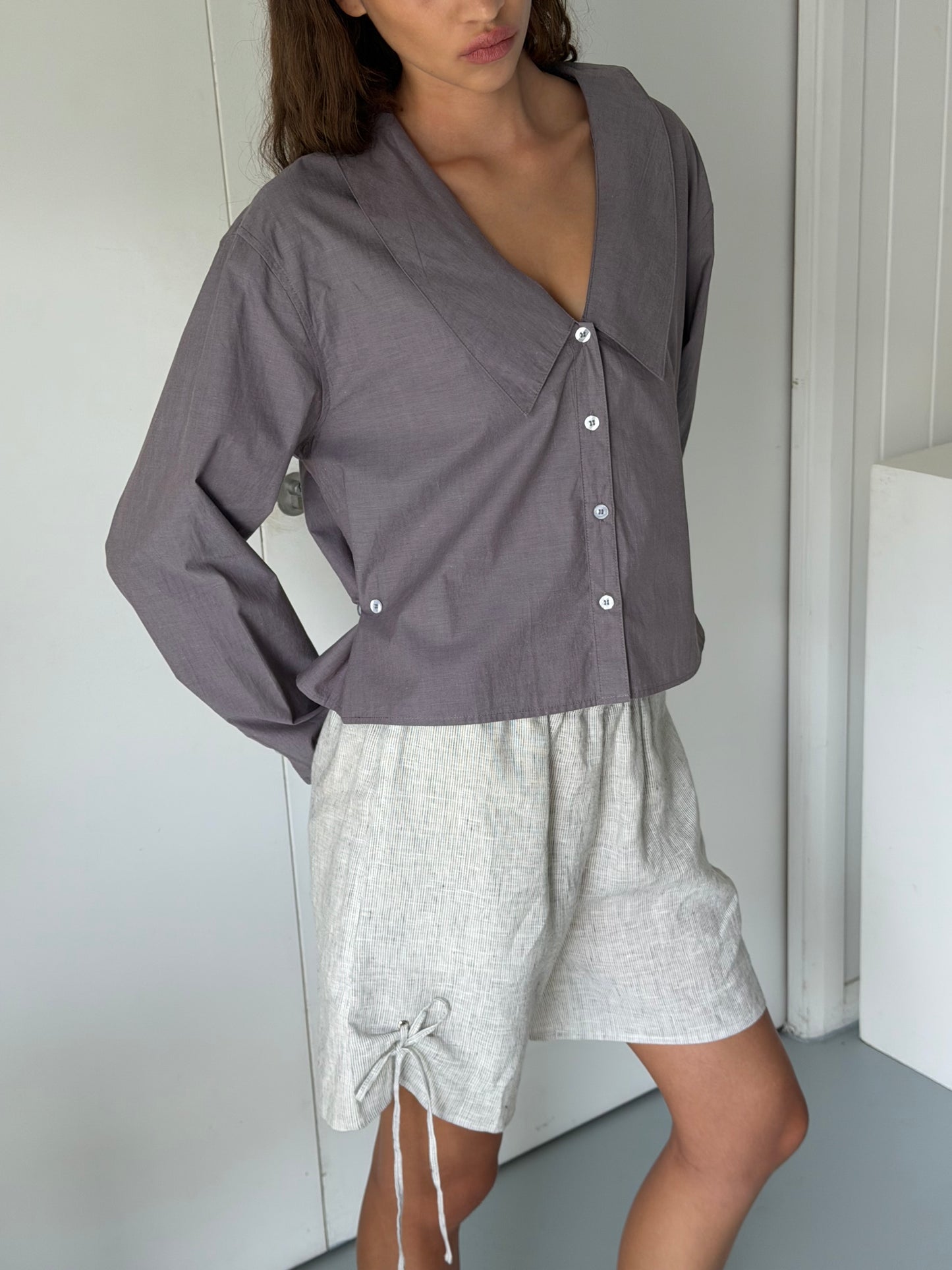 A person wears a loose Deiji Studios Waist Button Shirt in currant with a wide collar and light beige drawstring shorts, standing indoors with one hand on their hip.