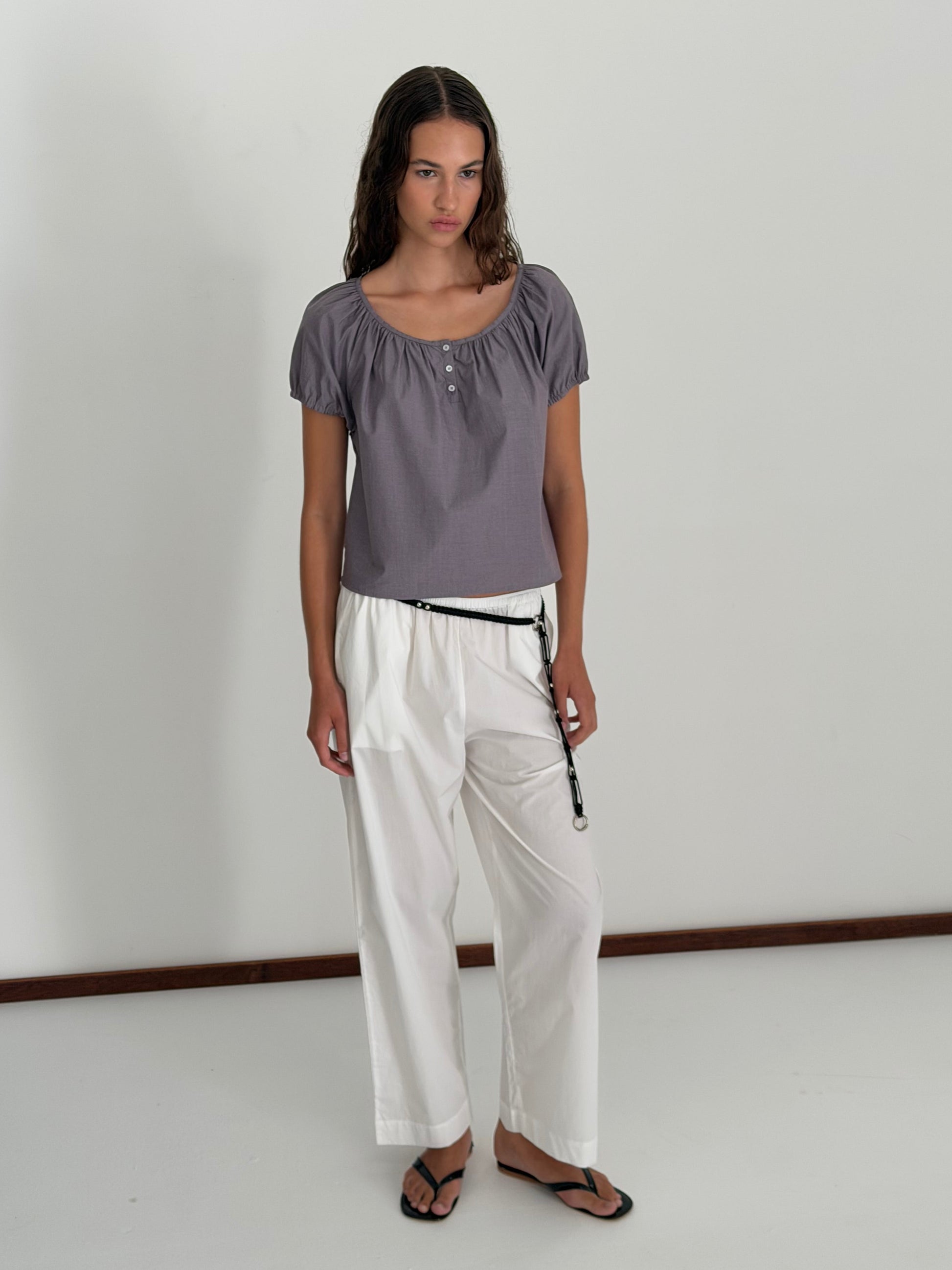 A woman with long wavy hair stands indoors against a white wall, wearing the Poplin T Shirt - Currant by Deiji Studios with loose white drawstring pants and black sandals, her expression neutral.
