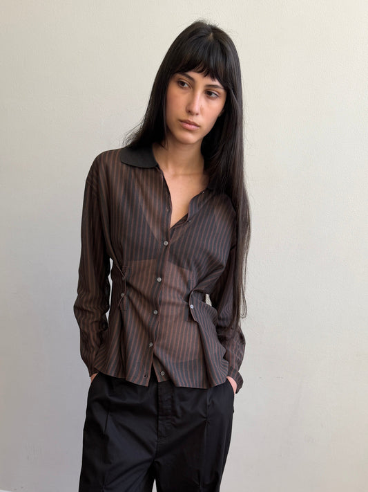 A woman with long dark hair wears the Knit Shirt - Brown Grey Stripe and black pants, standing against a plain light-colored wall with her hands in her pockets, looking slightly to the side.
