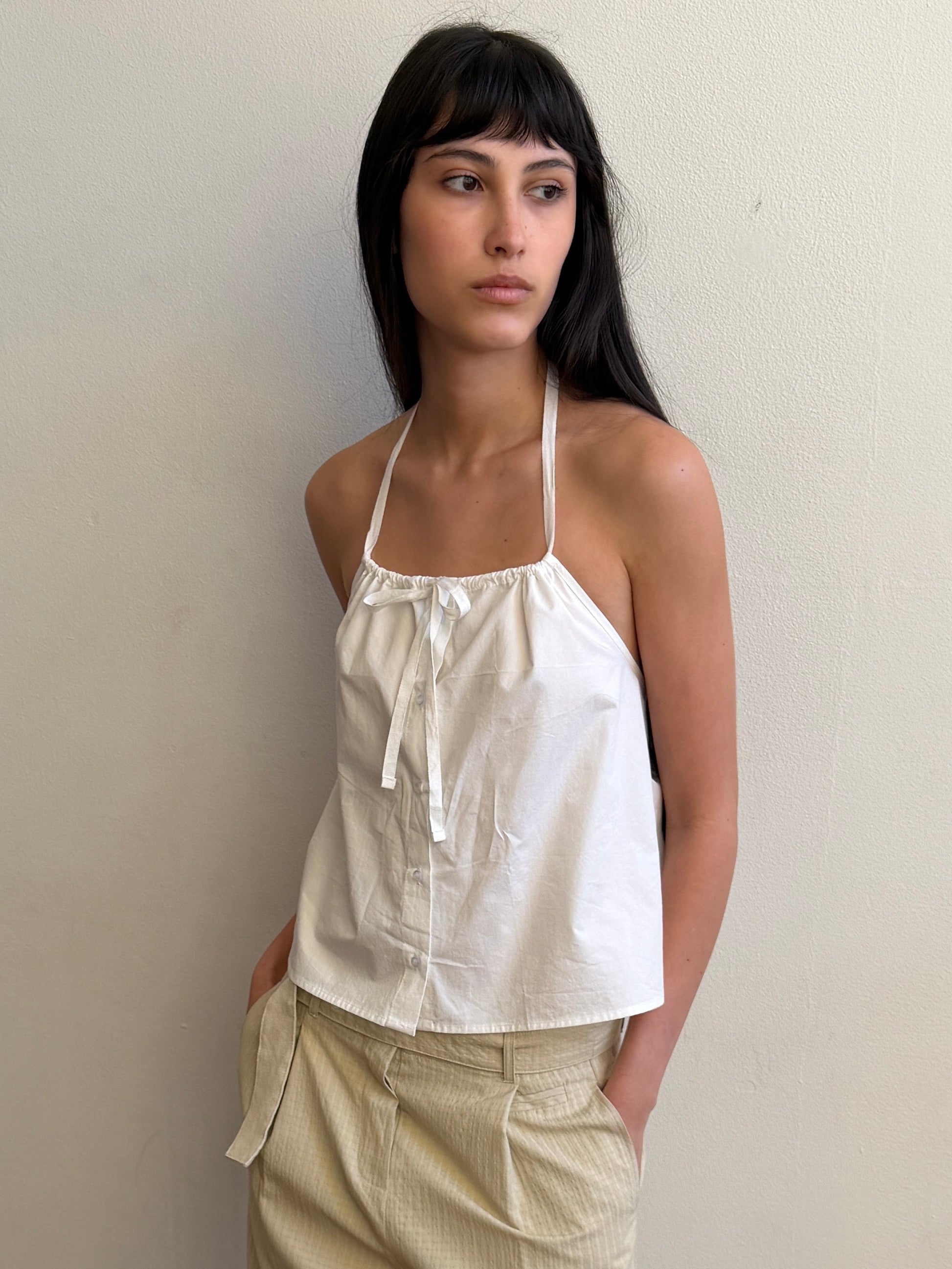 A woman with long dark hair wears the Simple Tie Top - White by Deiji Studios and beige pants, standing against a plain light-colored wall and looking to her left.