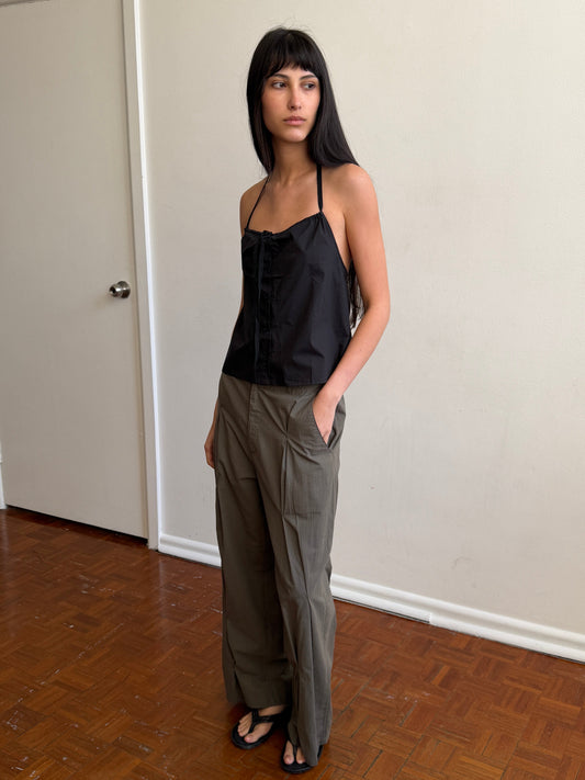 A woman with dark hair and a fringe stands in a room with a hand in her pocket, wearing the Multi Pleat Pant - Moss Stripe by Deiji Studios, moss-green fine-check wide-leg pleated trousers, with a black halter top and black sandals.