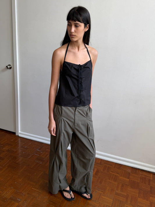 A woman with dark hair and a fringe stands in a room wearing the Multi Pleat Pant - Moss Stripe by Deiji Studios, moss-green fine-check wide-leg pleated trousers, with a black halter top and black sandals.