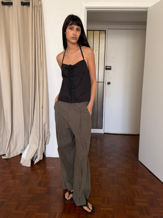 A woman with dark hair and a fringe stands in a room wearing the Simple Tie Top - Black by Deiji Studios, a black halter top with a gathered neckline and front tie, with khaki trousers and black sandals.