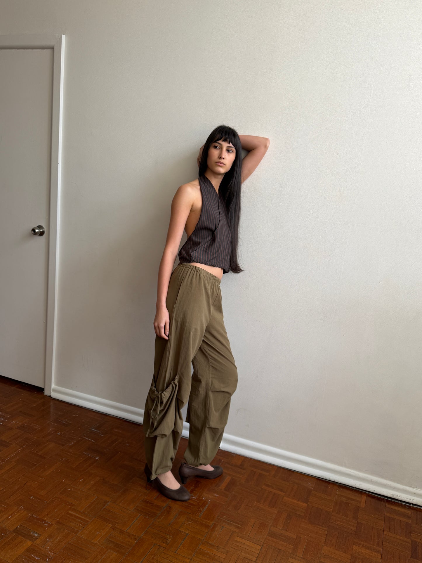 A person with long dark hair poses indoors against a plain, off-white wall, wearing the Wrap Halter Top in Brown Grey Stripe, paired with loose khaki pants and brown flats, with one arm raised behind their head.