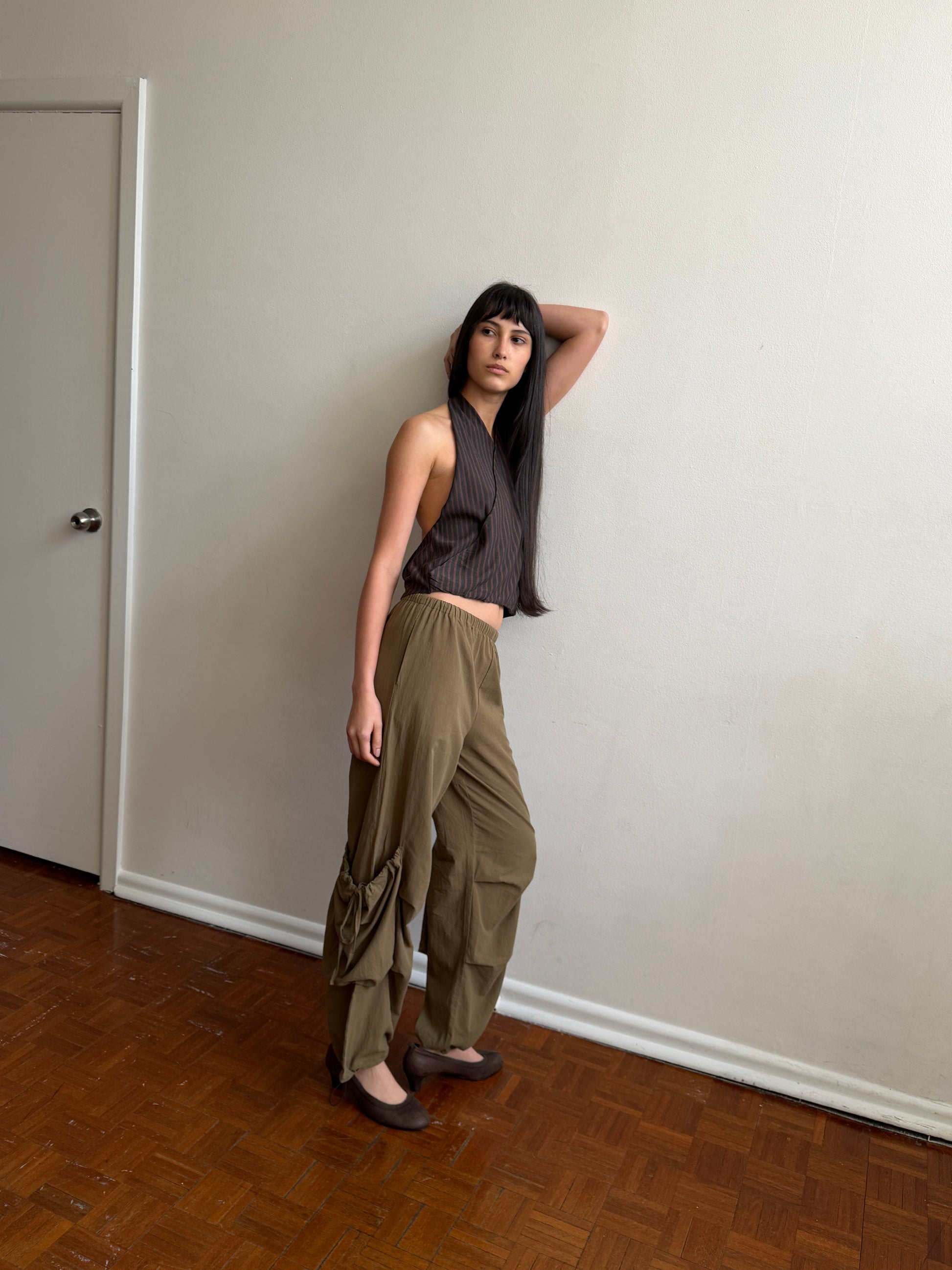 A person with long dark hair poses indoors against a plain, off-white wall, wearing the Wrap Halter Top in Brown Grey Stripe, paired with loose khaki pants and brown flats, with one arm raised behind their head.