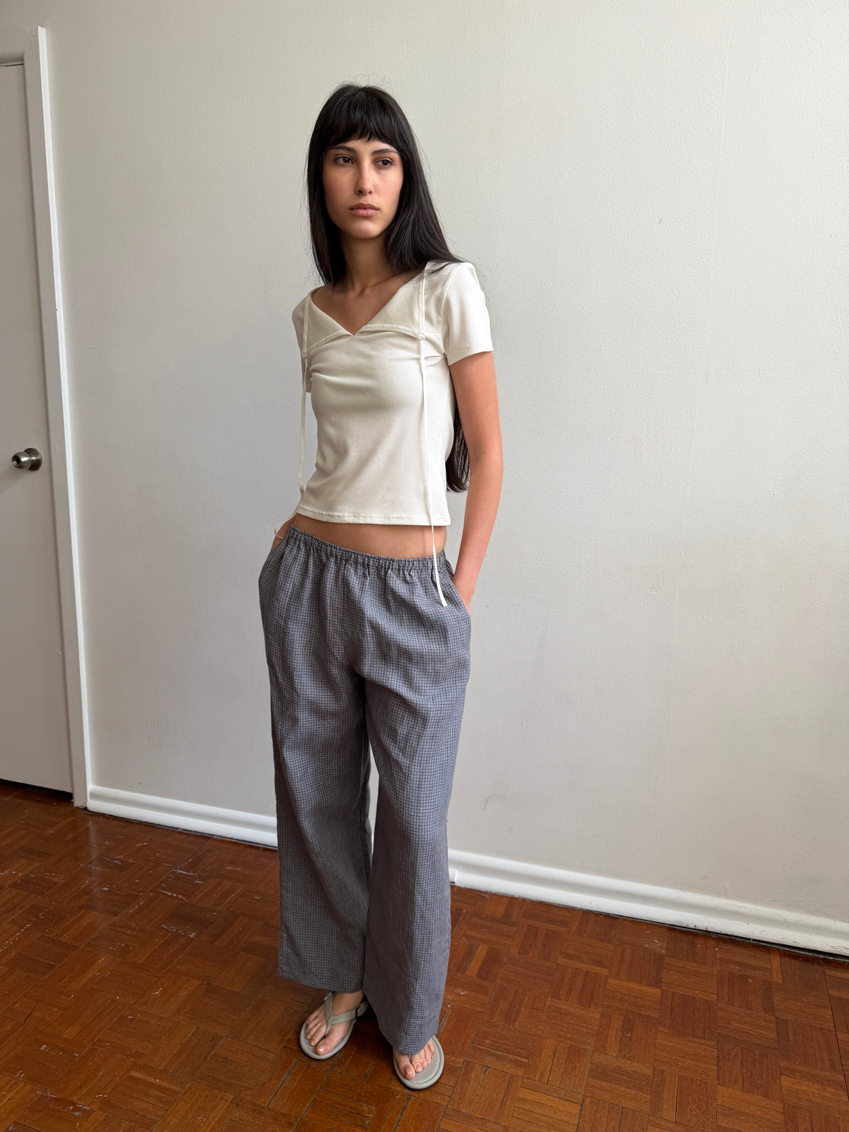 A woman with dark hair and a fringe stands by a door wearing the Linen Ease Trouser - Indigo Check by Deiji Studios, indigo micro-check wide-leg linen trousers, with a cream tie-back tee and grey flip-flops.