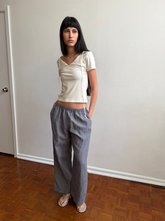 A woman with dark hair and a fringe stands by a door wearing the Linen Ease Trouser - Indigo Check by Deiji Studios, indigo micro-check wide-leg linen trousers, with a cream tie-back tee and grey flip-flops.