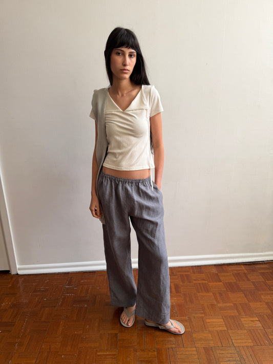 A woman with dark hair and a fringe stands in a room wearing the Linen Ease Trouser - Indigo Check by Deiji Studios, indigo micro-check wide-leg linen trousers, with a cream tie-back tee and grey flip-flops.
