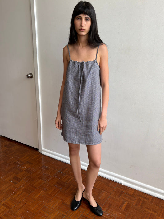 A woman with dark hair and a fringe stands in a room wearing the Weave Mini - Indigo Check by Deiji Studios, an indigo micro-check linen mini dress with thin straps and a drawstring front tie, with black flats.
