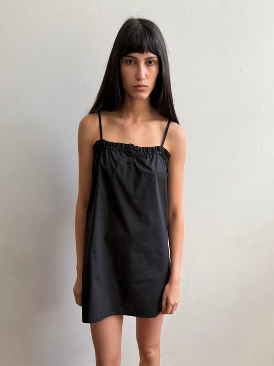 A woman with dark hair and a fringe stands against a wall wearing the Frill Placket Dress - Black by Deiji Studios, a black mini dress with thin straps and a gathered, buttoned frill placket at the neckline.