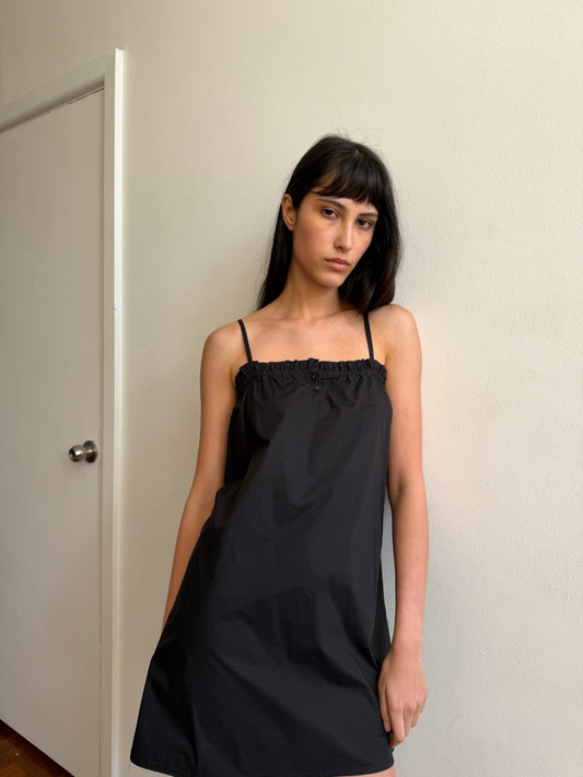 A woman with dark hair and a fringe stands against a wall wearing the Frill Placket Dress - Black by Deiji Studios, a black A-line mini dress with thin straps and a gathered, buttoned frill placket at the neckline.