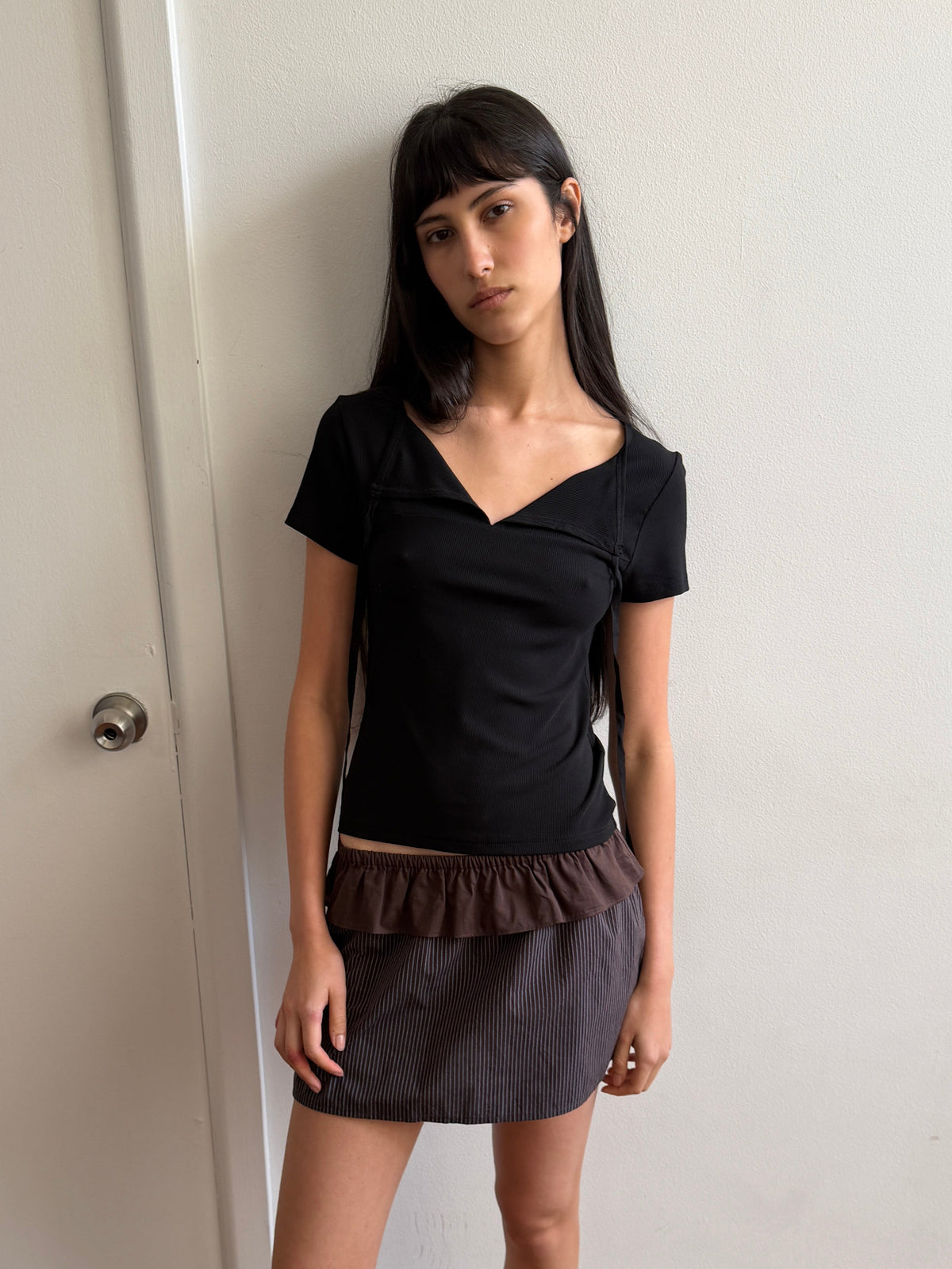 A woman with long dark hair stands indoors against a white wall, wearing a black short-sleeve top and the Flounce Layer Mini Skirt in Brown Blue Stripe, featuring a delicate flounce layer and an elasticated waistband, looking calmly at the camera.