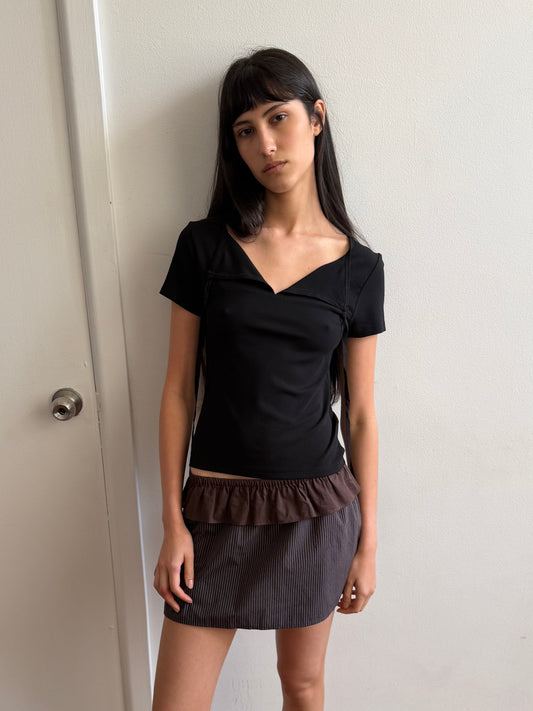 A woman with long dark hair stands indoors against a white wall, wearing a black short-sleeve top and the Flounce Layer Mini Skirt in Brown Blue Stripe, featuring a delicate flounce layer and an elasticated waistband, looking calmly at the camera.