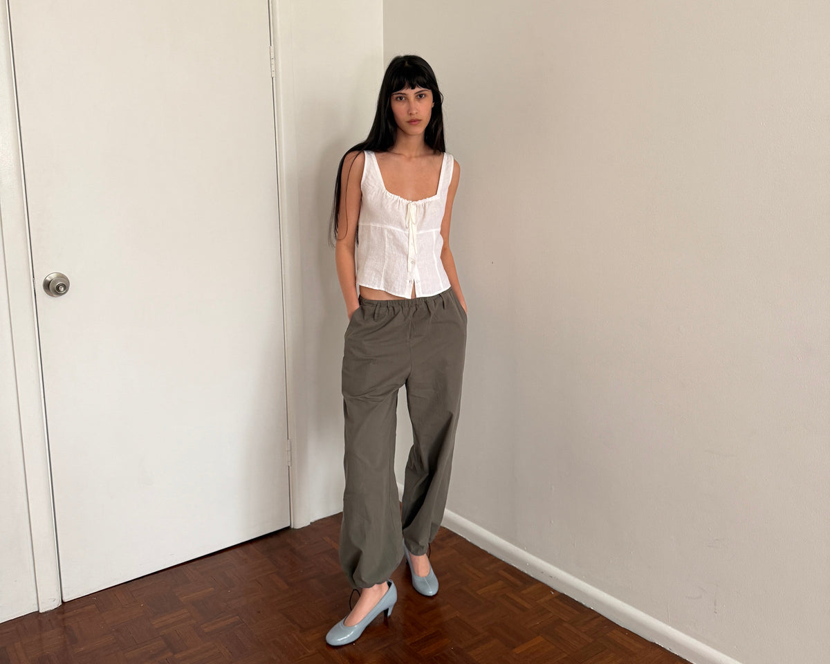 A woman with dark hair and a fringe stands in a room wearing the Relax Tie Top - White by Deiji Studios, a white sleeveless linen top with a drawstring tie neckline, with khaki trousers and pale blue heels.