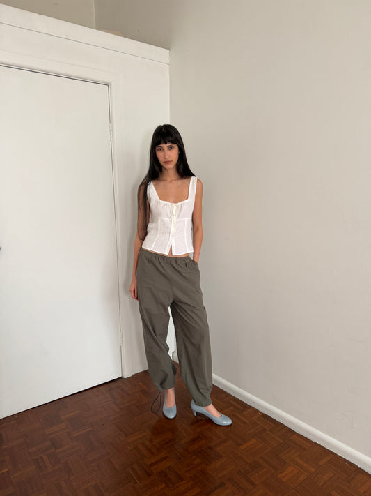 A woman with dark hair and a fringe stands against a wall wearing the Relax Tie Top - White by Deiji Studios, a white sleeveless linen top with a drawstring tie neckline, with khaki trousers and pale blue heels.