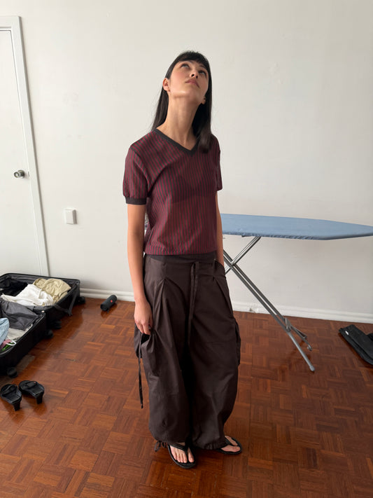 A woman with dark hair and a fringe stands in a room looking upward, wearing the Cross Neck Tee - Red Stripe by Deiji Studios, a fine red and charcoal striped knit tee, with dark brown drawstring wide-leg trousers.