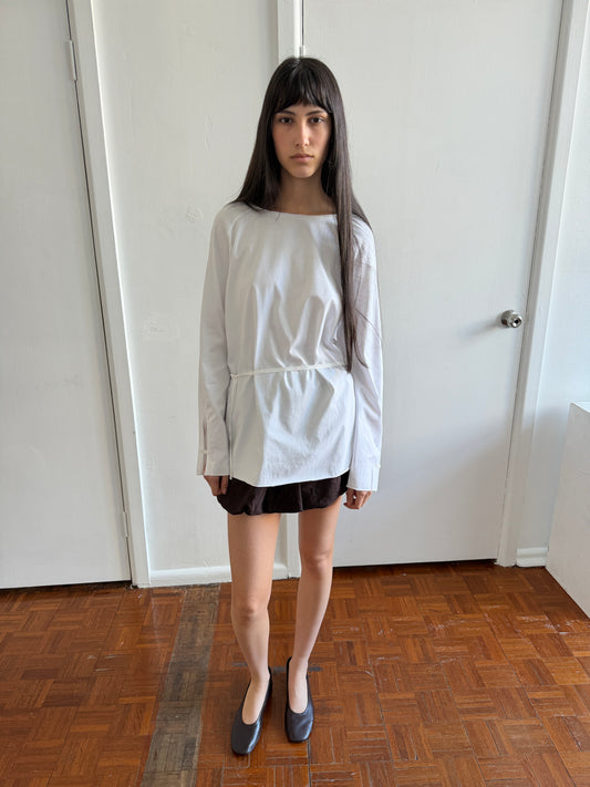 A woman with dark hair and a fringe stands in a room wearing the Wided Longsleeve - White by Deiji Studios, a white top with a wide neckline and drawstring hem, with a brown mini skirt and black flats.