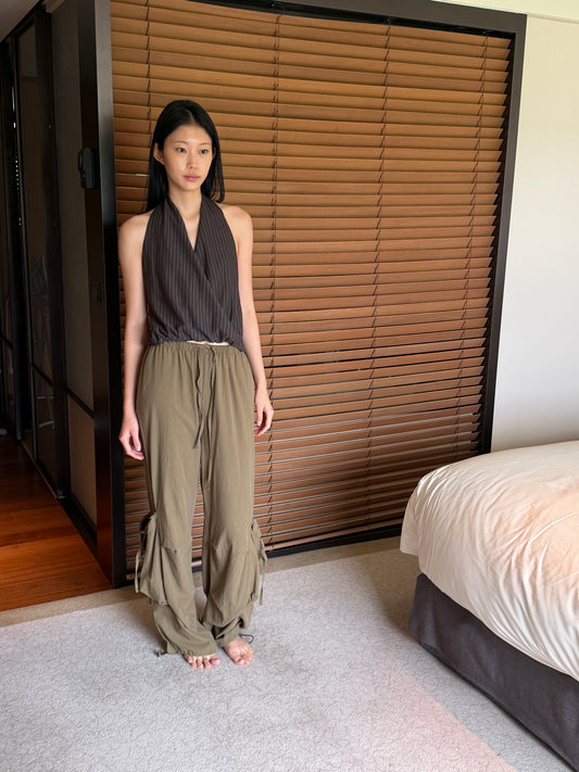A woman with long dark hair stands barefoot in a bedroom wearing the Drawcord Pocket Pant - Khaki by Deiji Studios, khaki wide-leg trousers with a drawcord waist, and a brown striped halter top.