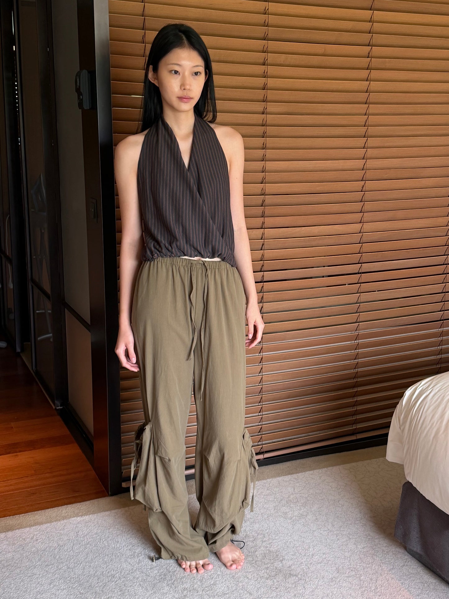 A woman with long dark hair stands barefoot in a bedroom wearing the Wrap Halter Top - Brown Grey Stripe by Deiji Studios, a sheer brown and grey striped halter top with a wrap front, and khaki trousers.