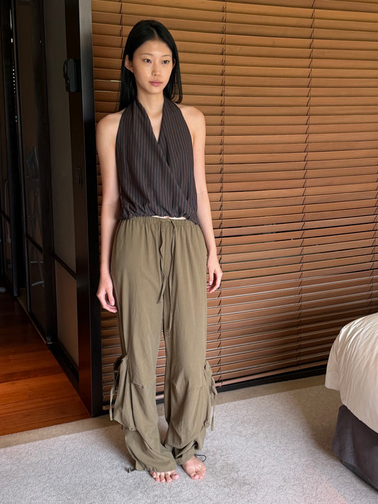 A woman with long dark hair stands barefoot in a bedroom wearing the Wrap Halter Top - Brown Grey Stripe by Deiji Studios, a sheer brown and grey striped halter top with a wrap front, and khaki trousers.