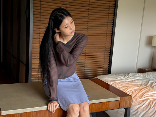 A woman with long dark hair sits on a bench in a wood-panelled room with a hand to her hair, wearing the Stripe Longsleeve - Brown Grey Stripe by Deiji Studios, a sheer brown and charcoal striped knit top, with a periwinkle midi skirt.