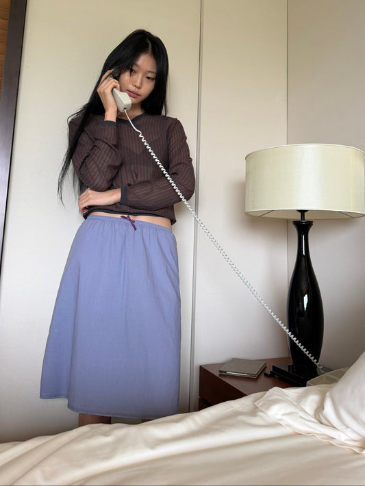 A woman reclines propped on one arm on a tatami-topped bench in a wood-panelled room, wearing a sheer brown striped long-sleeve top and the Basic Skirt - Thistle by Deiji Studios, a periwinkle cotton voile midi skirt with an elasticated waist.