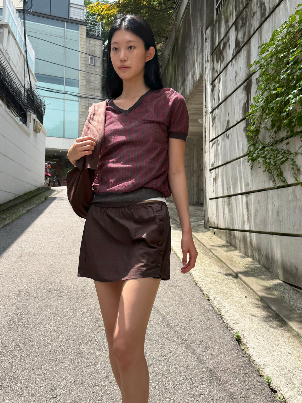A woman with long dark hair walks along a sunlit lane wearing the Cross Neck Tee - Red Stripe by Deiji Studios, a fine knit tee with red and charcoal vertical stripes and a V-neckline, paired with a brown mini skirt and a shoulder bag.