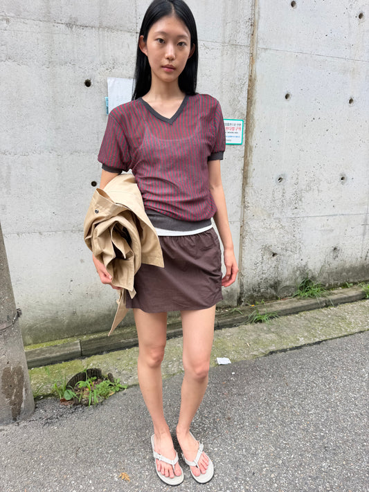 A woman with long dark hair stands against a concrete wall holding a beige jacket, wearing the Cross Neck Tee - Red Stripe by Deiji Studios, a fine red and charcoal striped knit tee, with a brown mini skirt.