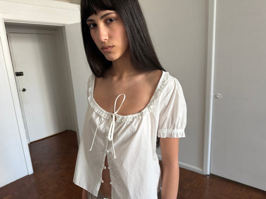 A cropped view of a woman with dark hair and a fringe wearing the Flounced Top - White by Deiji Studios, a white top with a frilled scoop neckline, front tie and puff sleeves, in a room.