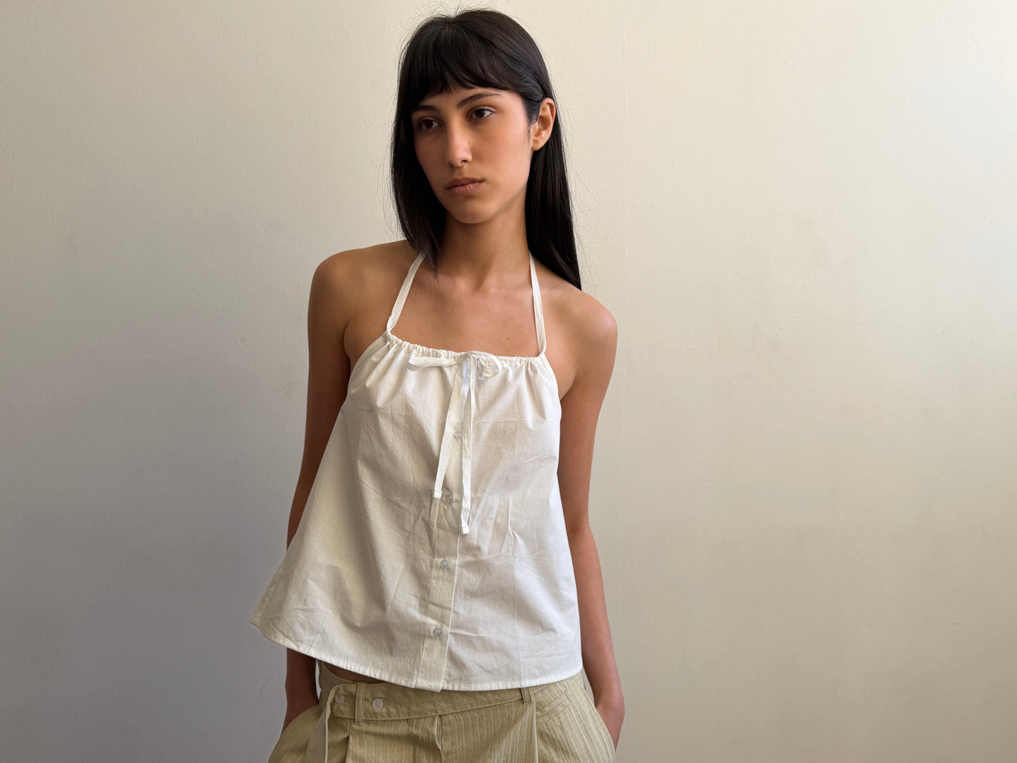 A person with long dark hair, wearing the Deiji Studios Simple Tie Top in white and light-colored pants, stands against a plain light-colored wall, looking slightly to the side with a neutral expression.