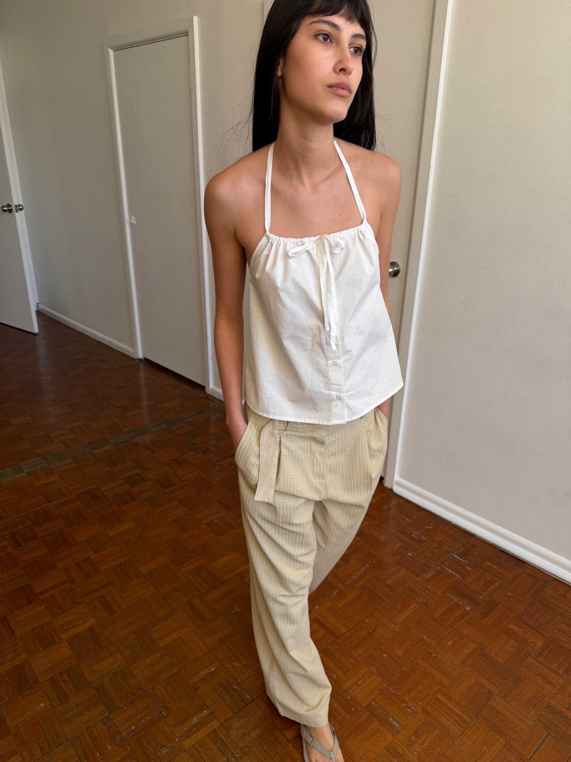 A woman wearing the Simple Tie Top - White from Deiji Studios and beige striped pants stands indoors on a wooden floor with her hands in her pockets, looking to the side.