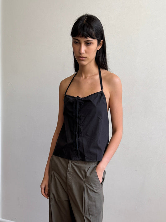 A woman with dark hair and a fringe stands in a room wearing the Simple Tie Top - Black by Deiji Studios, a black halter top with a gathered neckline and drawstring front tie, with khaki trousers.
