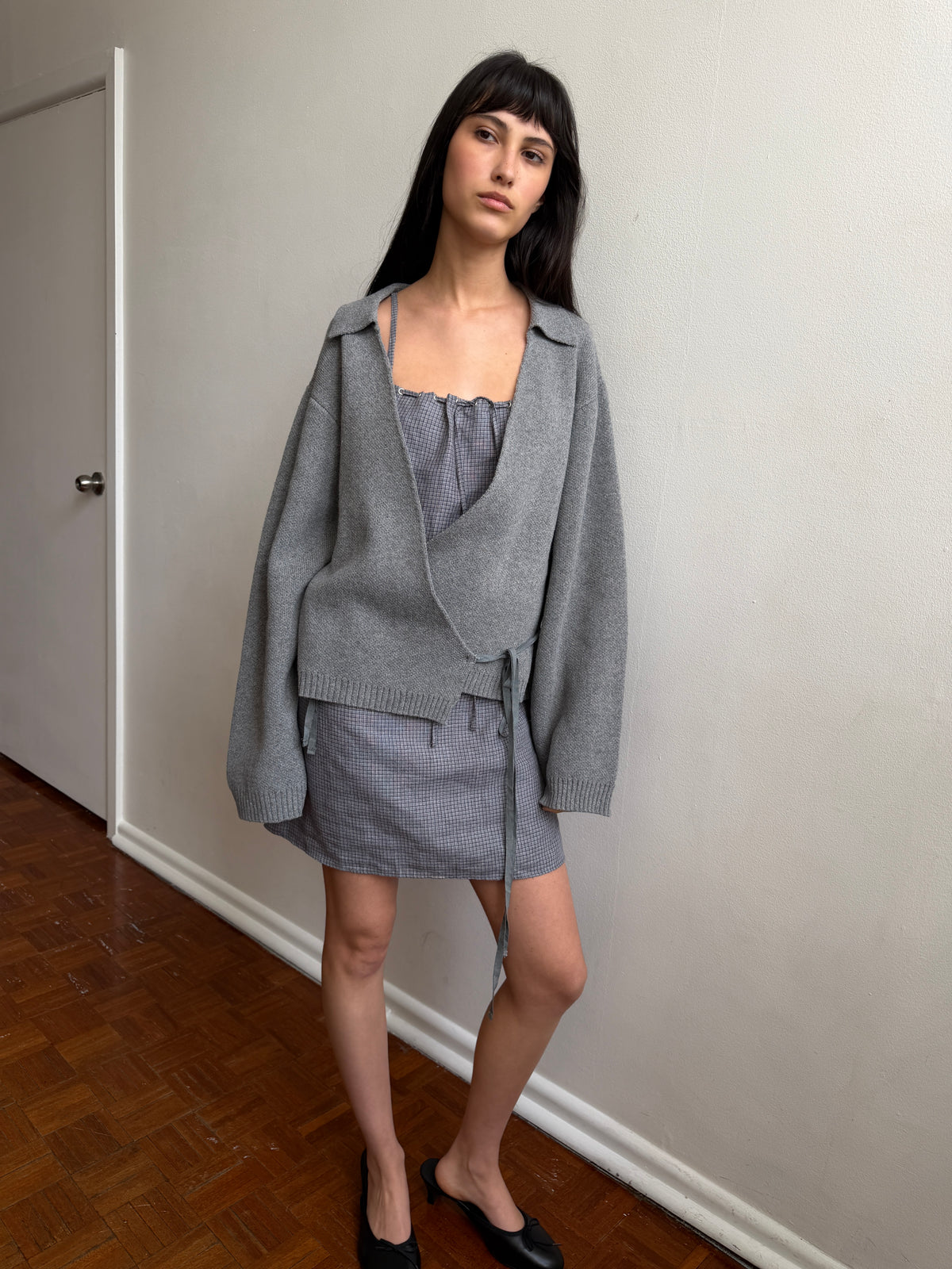 A woman with dark hair and a fringe stands in a room wearing the Wrap Cardigan - Grey by Deiji Studios, a grey knit wrap cardigan with a collar and waist tie, over a grey top and mini skirt with black flats.