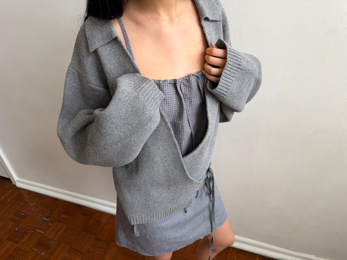 A cropped view of a woman wearing the Wrap Cardigan - Grey by Deiji Studios, a grey knit wrap cardigan with a collar, open over a grey top and mini skirt.